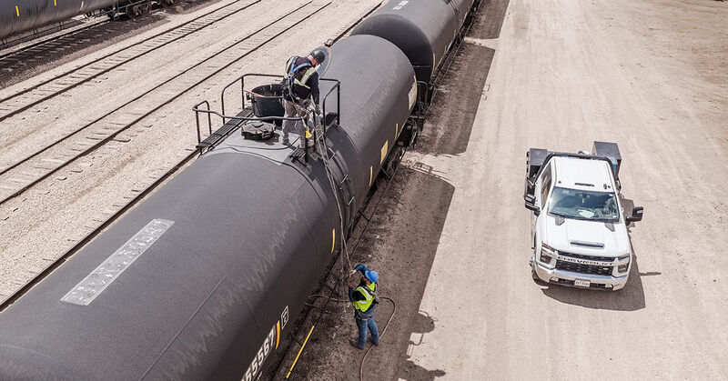 Two railway workers climb on and inspect a black tank car